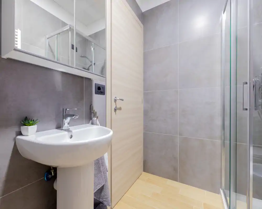 Salle de bain moderne et compacte avec lavabo sur colonne, miroir et douche vitrée ; carreaux gris et sol imitation bois pour un style propre et contemporain.