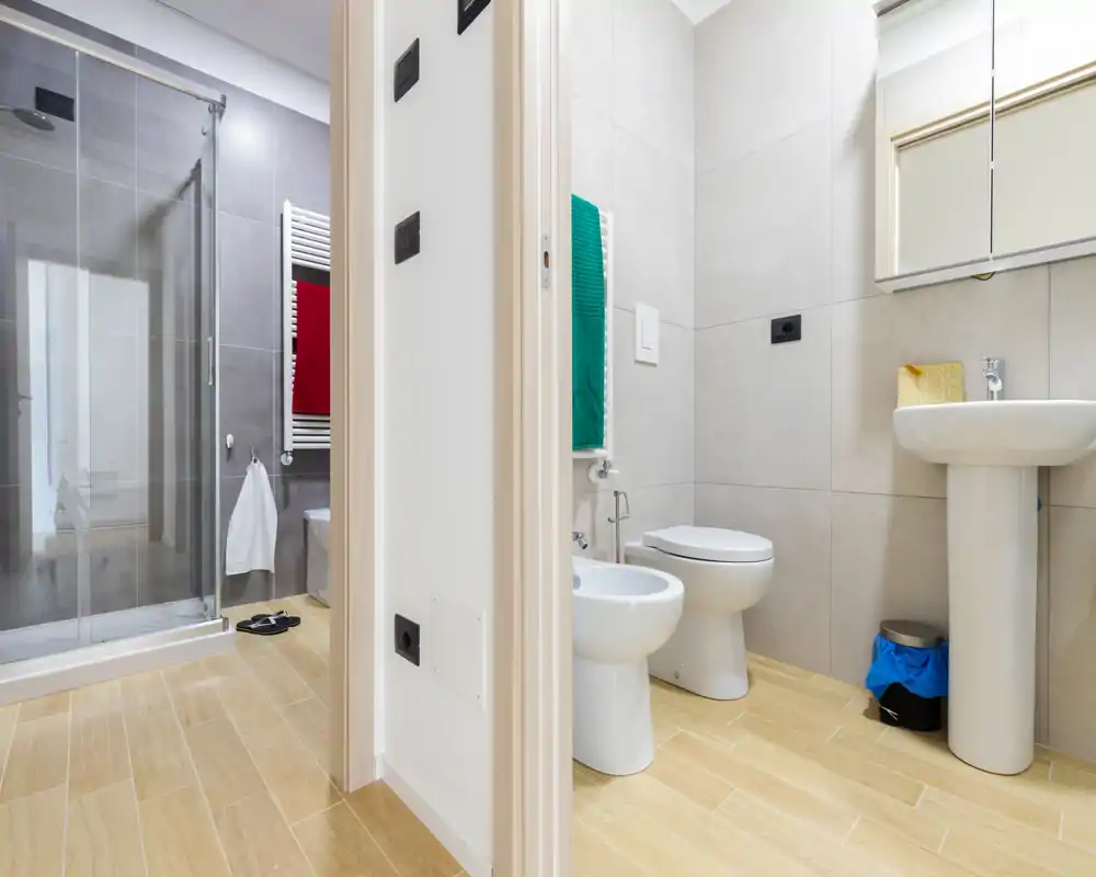 Salle de bains moderne avec lavabo sur colonne, bidet et WC, et une douche vitrée visible dans la zone adjacente ; carrelage clair et sol imitation bois pour un rendu propre et contemporain.