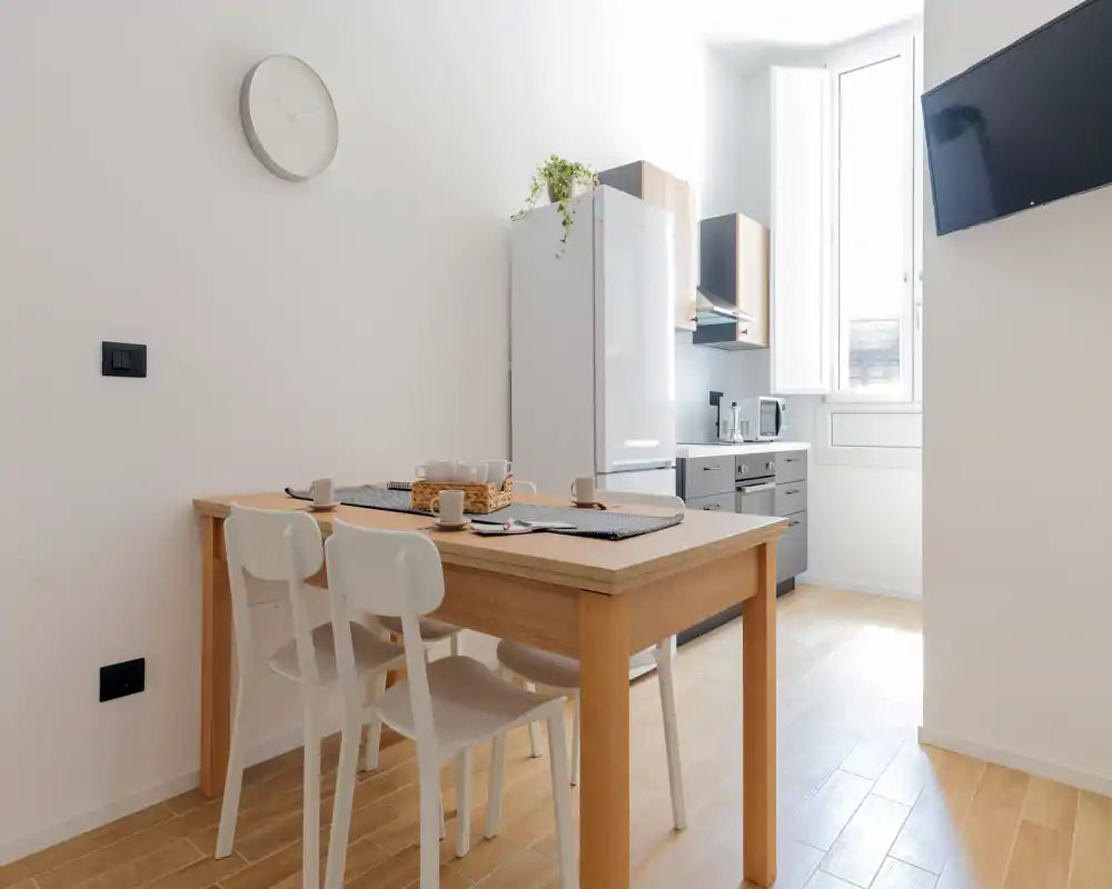 Coin repas moderne et lumineux avec une table en bois et quatre chaises blanches, proche d'une cuisine compacte et d'une grande fenêtre — idéal pour repas décontractés.