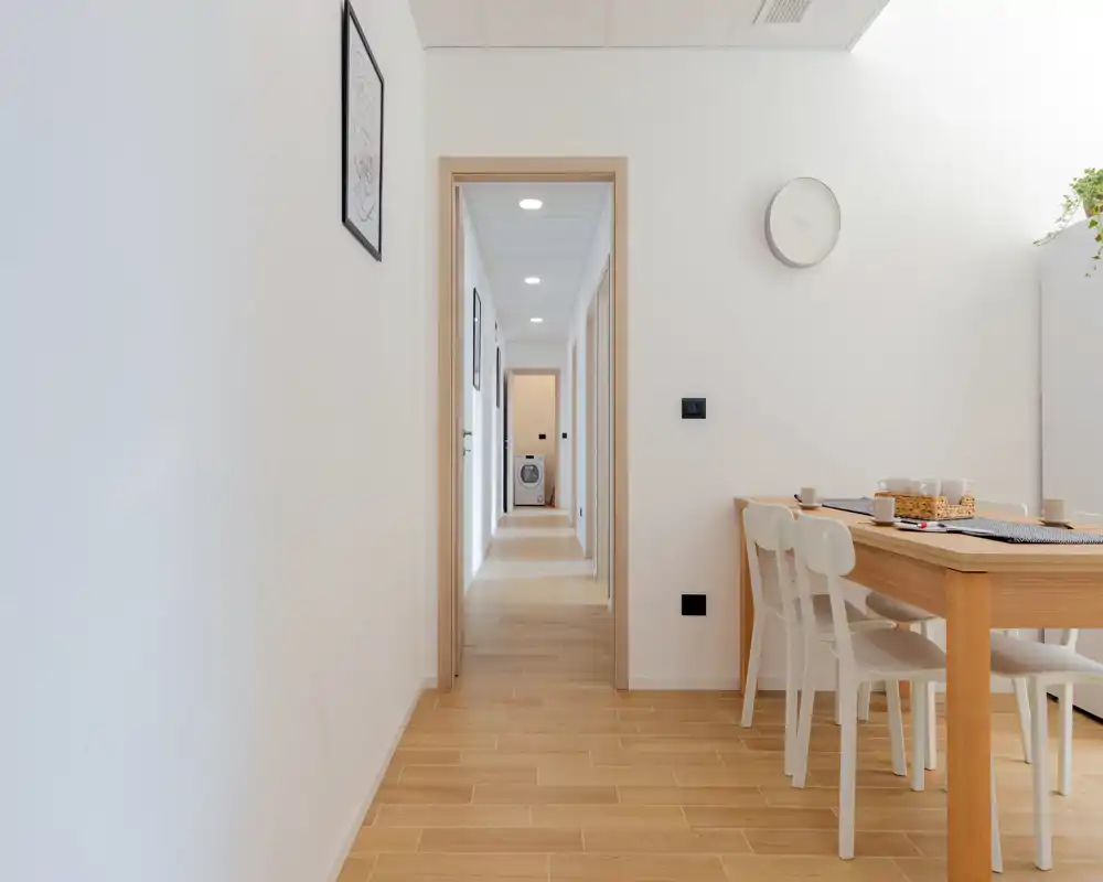 Coin repas lumineux et minimaliste avec une table en bois et des chaises blanches près d'un couloir ; lignes épurées et tons naturels pour une ambiance calme et aérée.