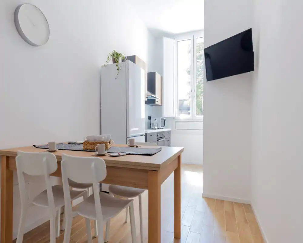 Coin repas moderne et lumineux ouvert sur une cuisine compacte. Table en bois pour quatre avec chaises blanches, sets de table et tasses, lumière naturelle abondante provenant d'une grande fenêtre et TV murale.