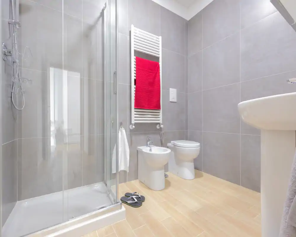 Salle de bain moderne et lumineuse avec cabine de douche en verre, bidet, WC, lavabo sur pied et radiateur porte-serviettes avec une serviette rouge.