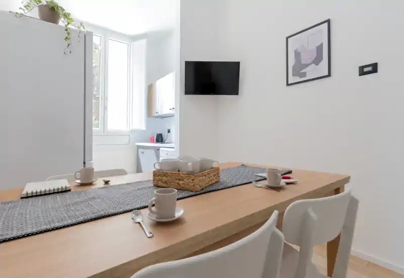 Zona pranzo luminosa e minimal con tavolo in legno apparecchiato per il caffè, sedie bianche moderne e luce naturale da una grande finestra che crea un’atmosfera accogliente.
