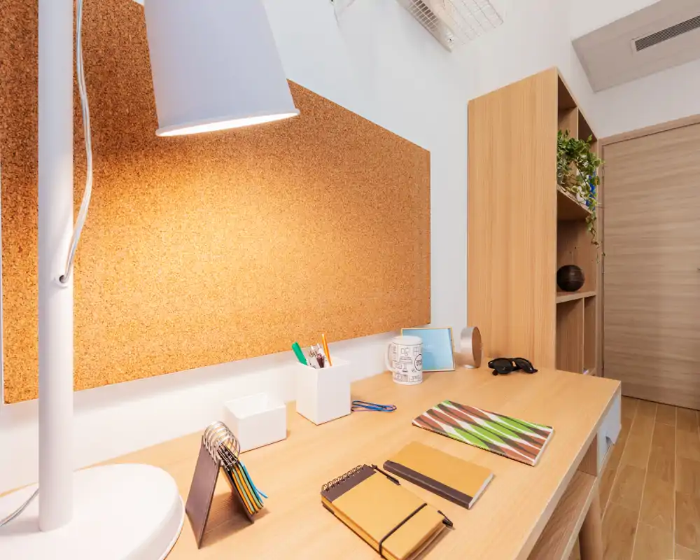 Espacio de estudio luminoso y nítido dentro del dormitorio, con escritorio de madera, material de papelería y un gran corcho; la imagen destaca la zona de trabajo más que la cama.