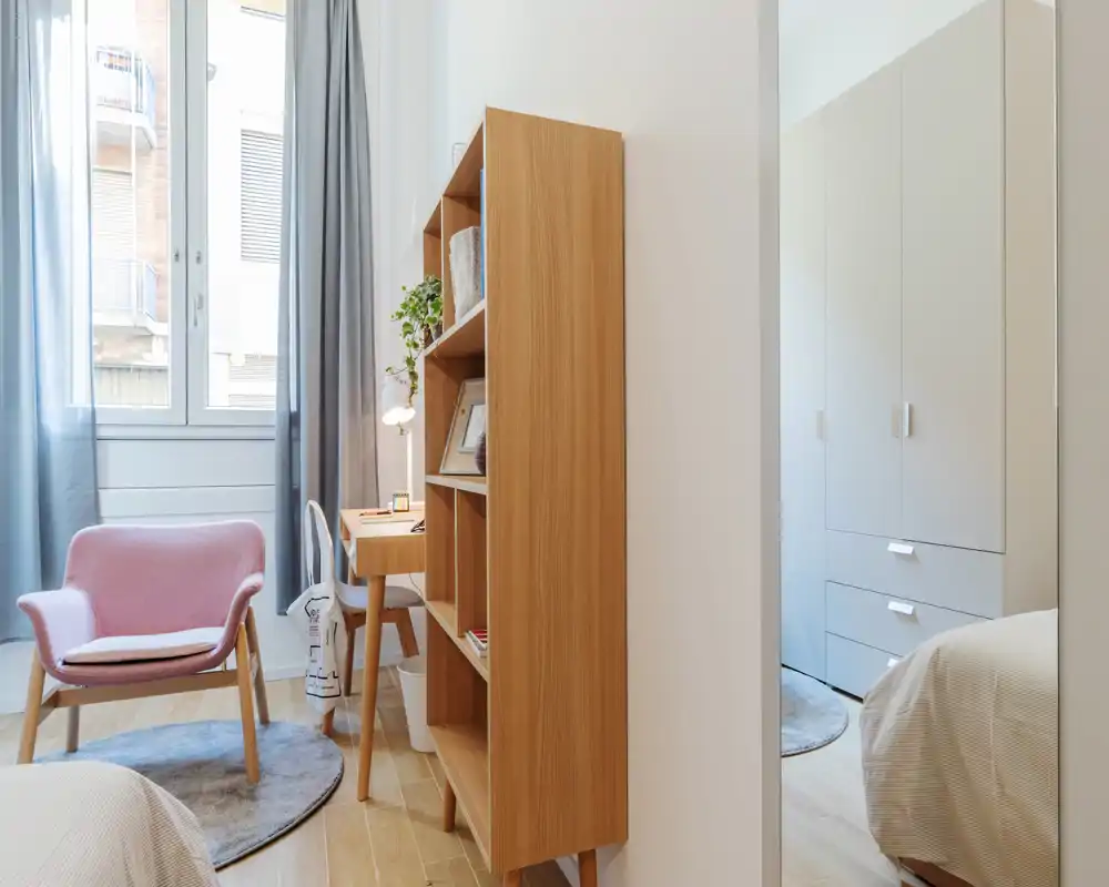 Rincón de dormitorio luminoso y acogedor con el borde de la cama visible, estantería de madera, butaca rosa y un pequeño escritorio junto a la ventana—luz natural y tonos cálidos que invitan.