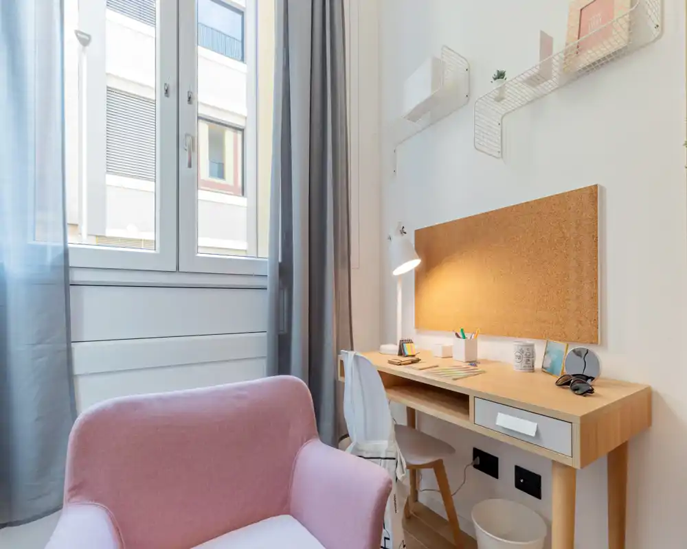 Rincón luminoso de salón/estudio con sillón rosa y escritorio de madera junto a la ventana. Perfecto como espacio acogedor para leer o trabajar con luz natural.