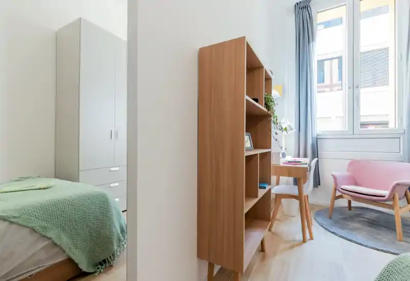 Bright, modern bedroom corner with a visible bed draped in a green knit throw, wardrobe, wooden shelving, a small desk by the window and a cozy pink armchair.