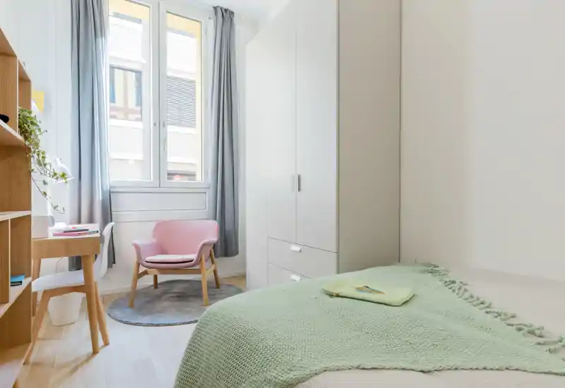 Sunny, compact bedroom with a neatly made bed draped in a soft green throw, a wardrobe, desk and a cozy pink armchair beside a large window.