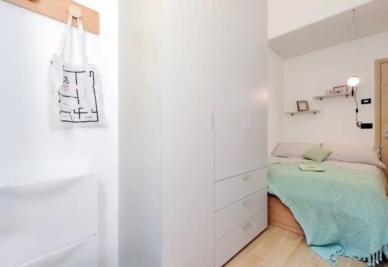Camera da letto compatta con armadio integrato e letto accogliente con morbide texture verdi; arredo minimal, illuminazione luminosa e mensole sul comodino per un ambiente ordinato.