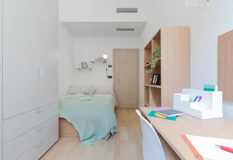 Bright, compact bedroom with a neatly made single bed dressed in a mint throw, built-in wardrobe, wooden shelving and a study desk — ideal for a student or short-term rental.