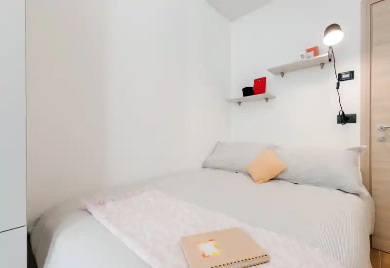 Dormitorio luminoso y minimalista con cama doble bien hecha, almohadas suaves y una pequeña manta. Estantes en la pared y una lámpara de noche añaden comodidad y funcionalidad.