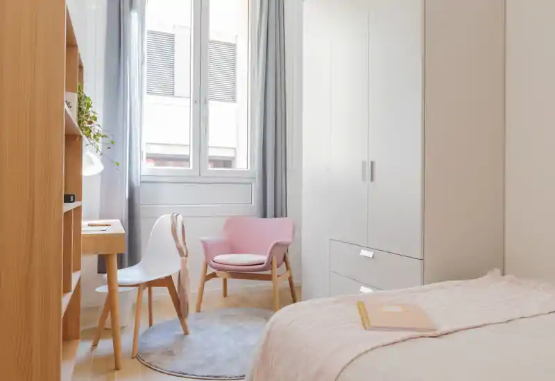 Dormitorio luminoso de estilo suave, con butaca rosa acogedora, cama arreglada con ropa de cama en tonos pastel, mobiliario de madera natural y una gran ventana que aporta mucha luz.