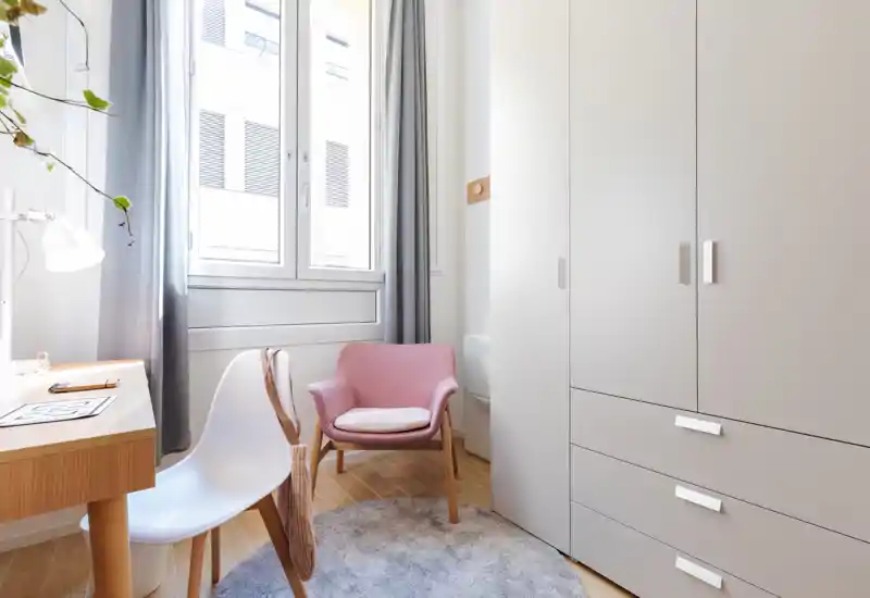 Bright, compact sitting area with a pink accent armchair, modern white chair, desk and large wardrobe next to a tall window allowing natural light.