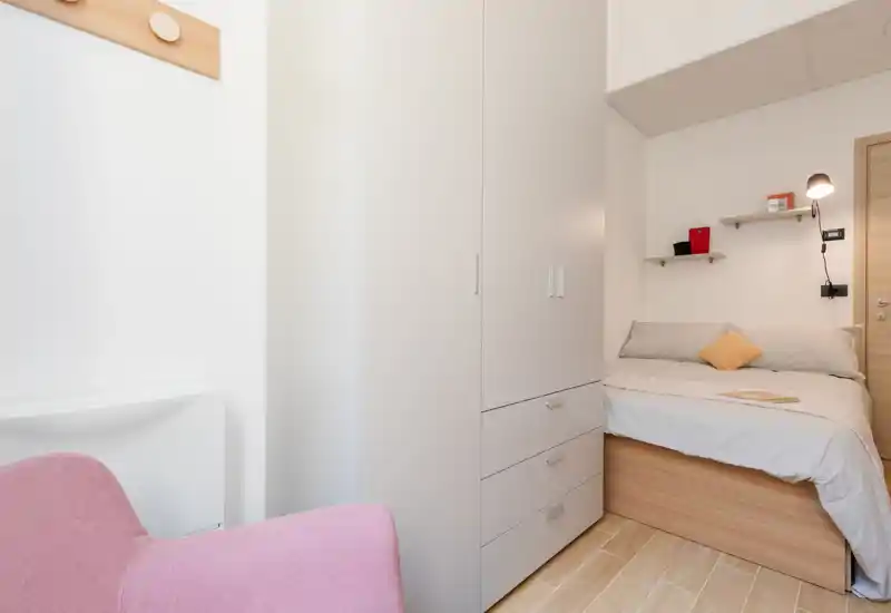 Dormitorio compacto y luminoso con una cama individual de madera, ropa de cama gris y un cojín mostaza. Un armario alto blanco y pequeñas estanterías en la pared maximizan el almacenamiento en este espacio ordenado y bien iluminado.