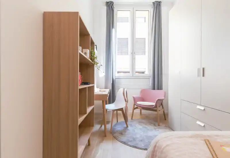 Dormitorio moderno y luminoso con toques en tonos pastel: cama parcialmente visible, estantería de madera, escritorio y un acogedor sillón rosa junto a la ventana que aporta luz natural.