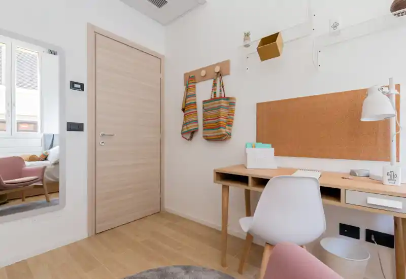 Small bedroom corner with a neat wooden desk, cork noticeboard and a white chair; a mirror at left reflects the adjacent bed and armchair. Bright, warm decor with striped hanging bags and soft wood flooring.