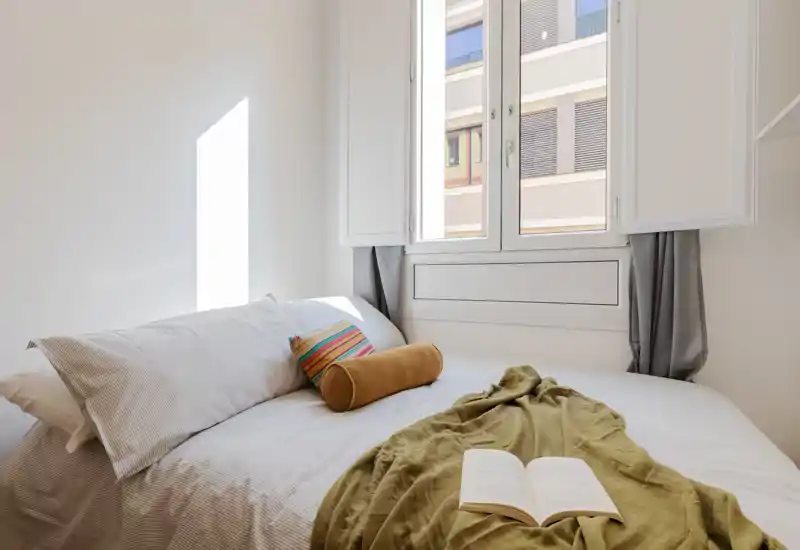 Bright, cozy bedroom with a neatly made double bed, soft linens, decorative cushions and natural light from a large window.
