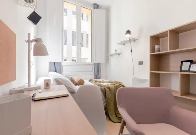 Bright, modern single bedroom with a desk and comfortable armchair. Natural light fills the room from a large window, highlighting a neatly made bed with cozy textiles and minimal, stylish shelving.