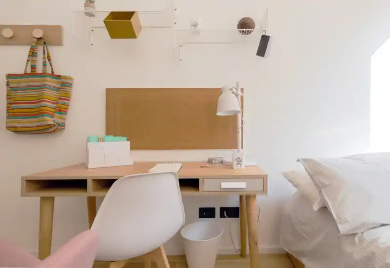 Bright, tidy bedroom corner with a desk and a partially visible bed. Natural light, warm wood tones and simple decor create a calm workspace next to the sleeping area.