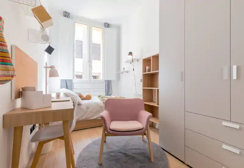 Bright, compact bedroom with a single bed under a large window, cozy textiles and a pink accent armchair. Built-in wardrobe and small desk create a functional layout ideal for a student or single occupant.