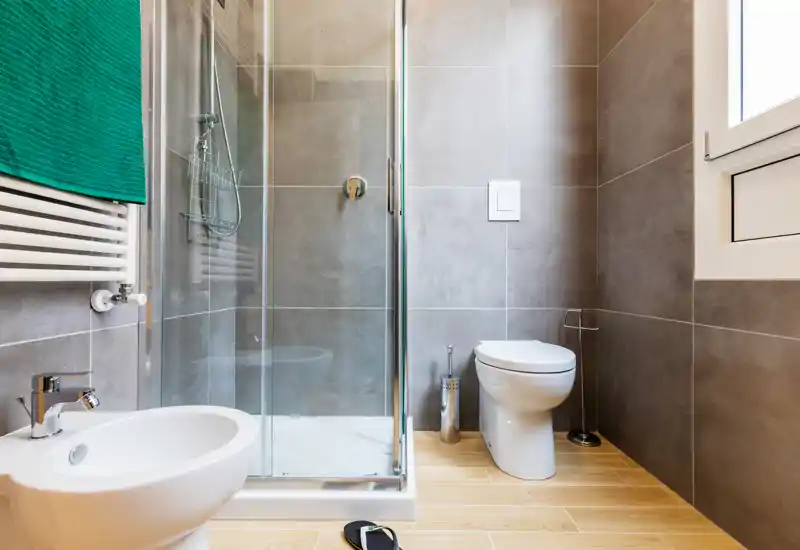 Modern bathroom with walk-in glass shower, wall-mounted tiles and a white toilet; light wood-look floor and a towel radiator visible on the left.