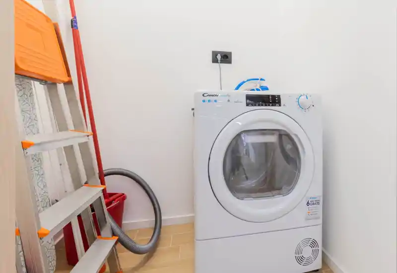 Small utility/laundry corner with a front-loading washing machine, stepladder and cleaning tools; bright, functional space suitable for practical use.