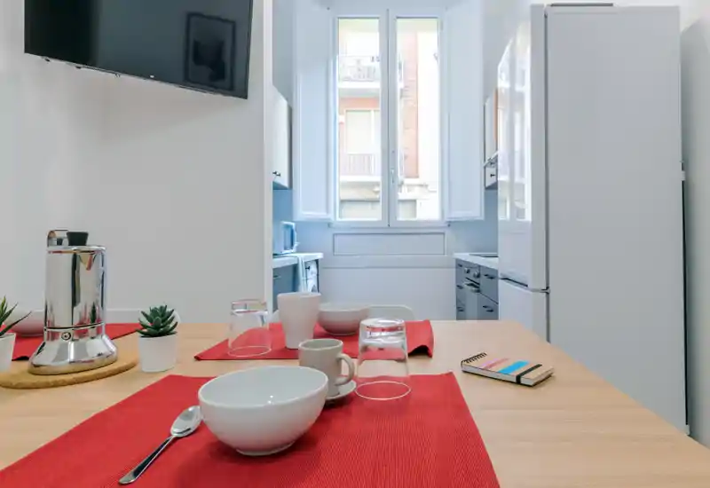 Bright, modern kitchen with a wooden dining surface set for breakfast: cups, bowls, glasses and red placemats. Natural light from a large window illuminates the tidy layout and appliances.