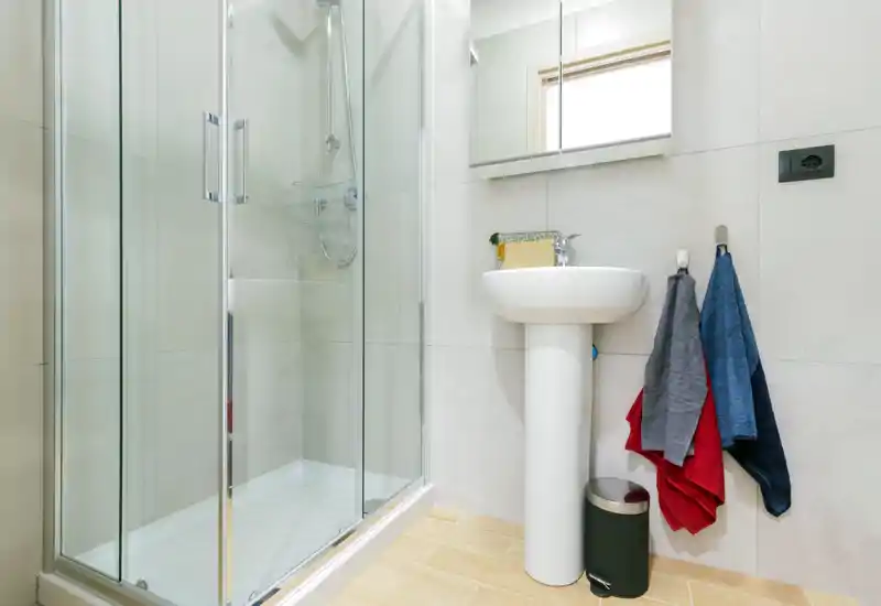 Bright, modern bathroom with a glass-enclosed shower, pedestal sink and wall-mounted mirror cabinet. Light tiles and wooden-effect floor create a clean, fresh look.
