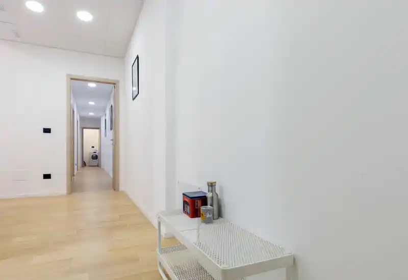 Bright, minimal hallway/entrance with light wood flooring, white walls and simple shelving — clean modern entry area with recessed ceiling lights and a view down a corridor.