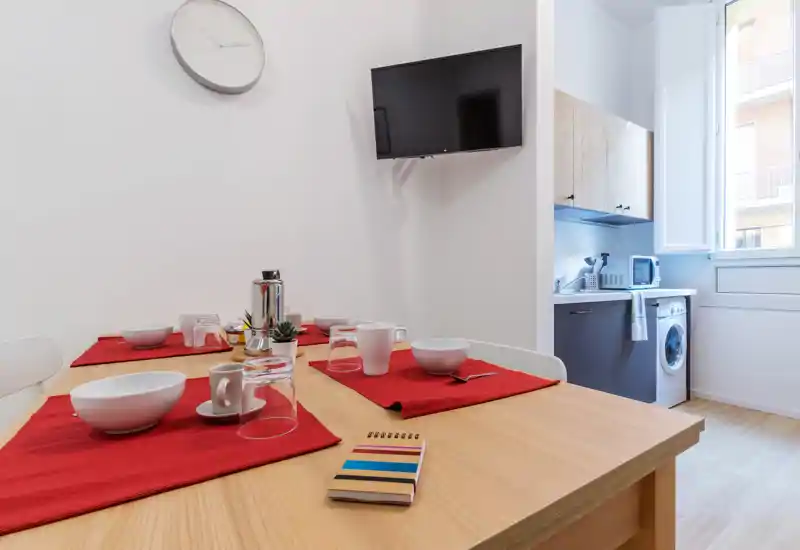 Bright, modern dining area with a wooden table set for meals, red placemats and tableware; an adjacent compact kitchenette and a window provide natural light.