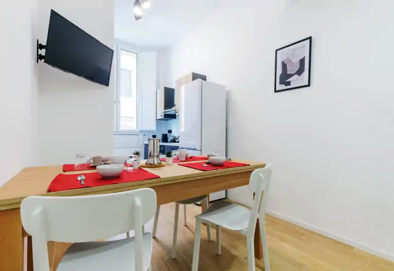 Bright, minimal dining area with a wooden table set for four, red placemats and simple white chairs adjacent to a compact open kitchen.