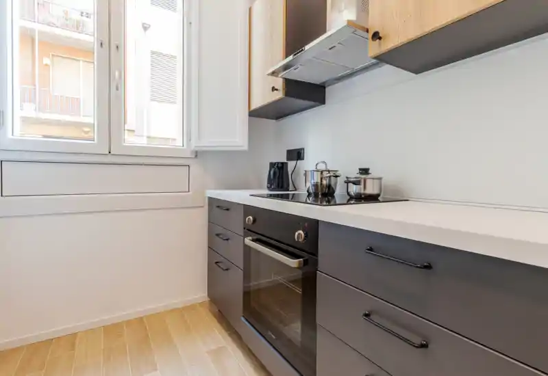 Bright modern kitchen with a built-in oven, induction hob and wooden floor. Natural light from a large window highlights the clean white countertop and contemporary cabinetry.