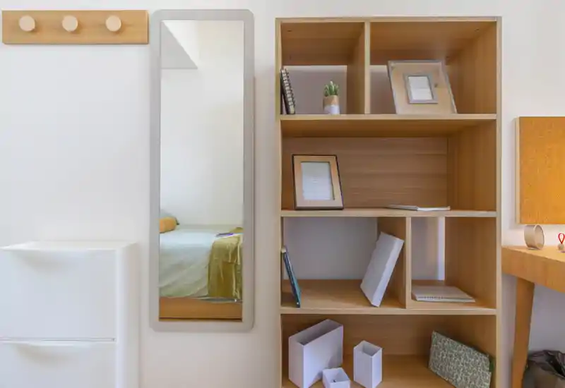A tidy bedroom corner seen through a wall mirror and adjacent shelving: a bed is partially visible in the mirror, with light linens and a neat wooden cubed bookcase at right.