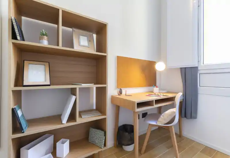Bright, compact workspace with a wooden desk, corkboard and shelving — ideal for study or remote work. Natural light from the window and a desk lamp create a pleasant, organized atmosphere.