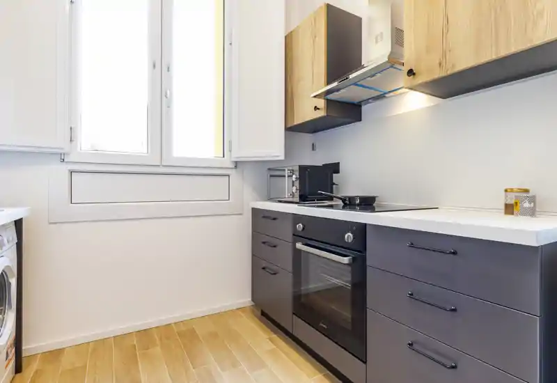 Bright modern kitchen with built-in oven, induction cooktop and wood-accent cabinets. Natural light from a large window and clean countertops create a fresh, functional space.