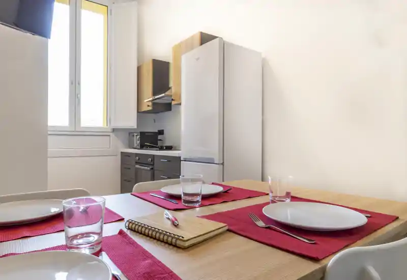 Bright, neatly set dining area with a wooden table laid for four, red placemats and simple modern tableware near an open window and a compact kitchen in the background.