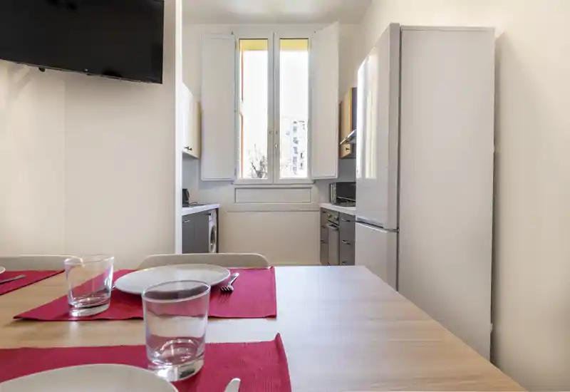 Bright, compact kitchen with a dining table set with red placemats, a refrigerator, and natural light from a central window.