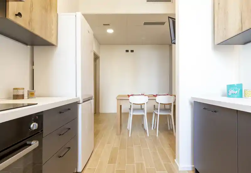 Modern galley kitchen opening onto a small dining area; light wood floors, white countertops and integrated appliances create a bright, tidy space ideal for short-term rentals.