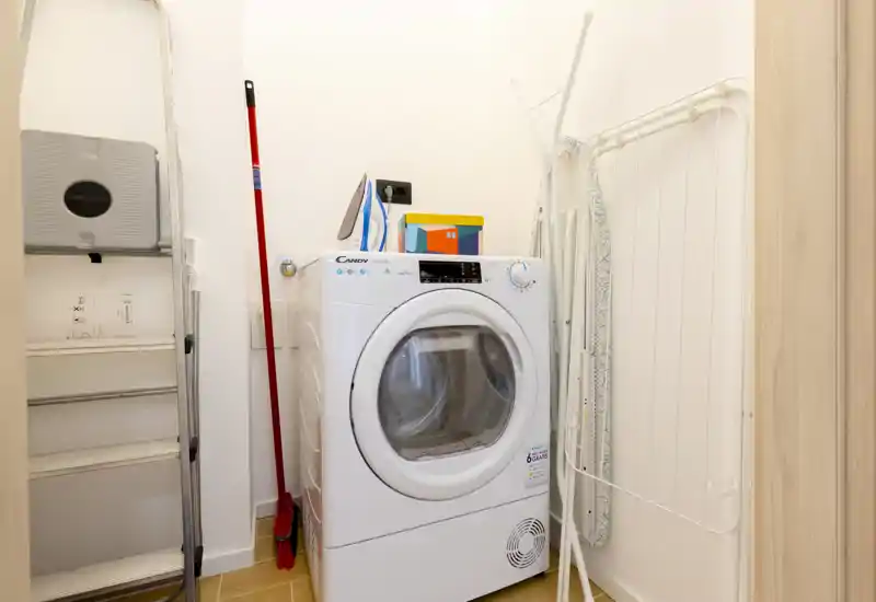 Small utility/laundry closet with a front-loading washer/dryer, ironing board, ladder and cleaning tools. Bright, compact space ideal for household chores.