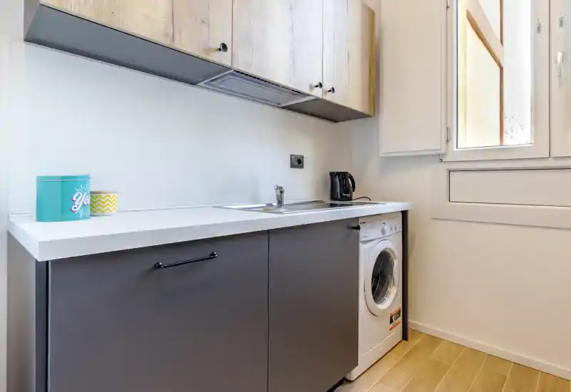 Compact modern kitchenette with a white countertop, integrated sink, upper wood-effect cabinets and a washing machine under the counter; bright natural light from the window.