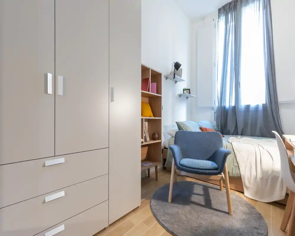 Camera da letto luminosa e ben arredata con letto confortevole, poltrona per la lettura e armadio; luce naturale dalle finestre e tessuti morbidi che creano calore.