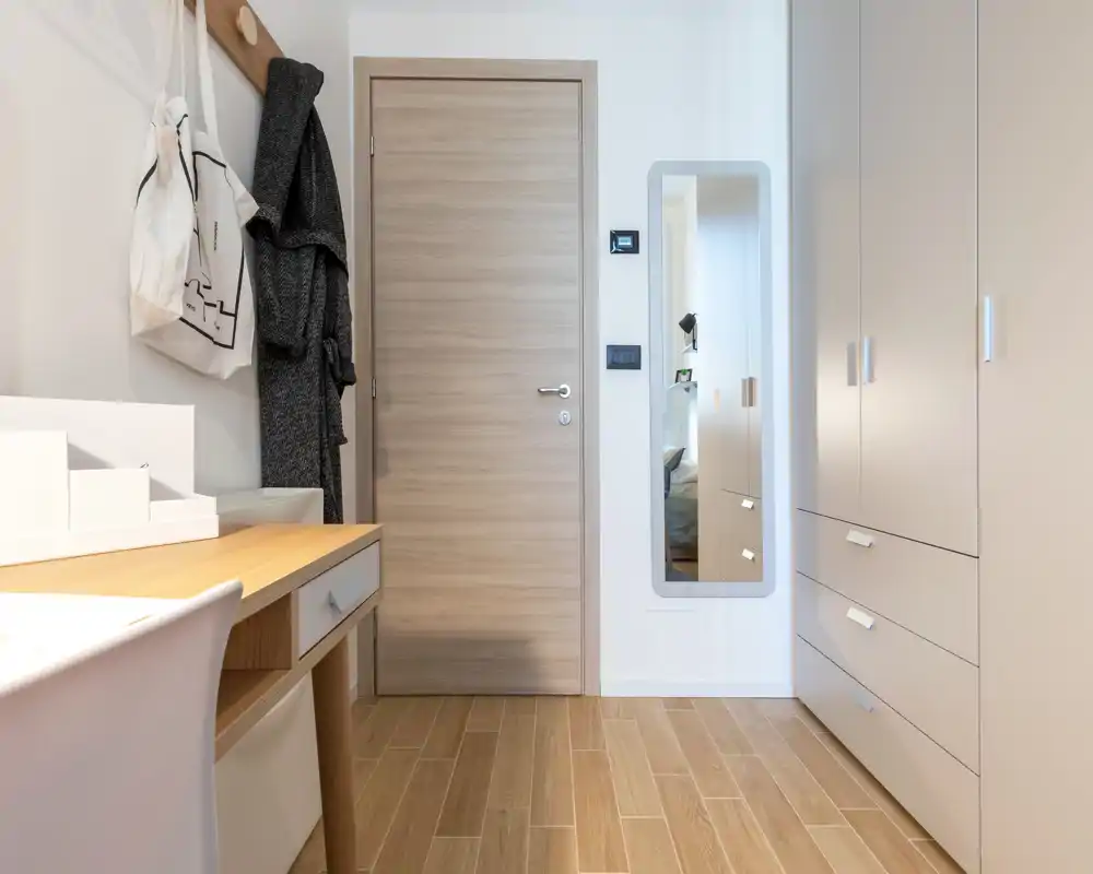 Ingresso ben illuminato con porta in legno, appendiabiti a parete con giacca e borsa, specchio a figura intera e armadi a cassetti integrati — pulito e moderno.