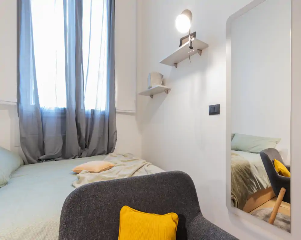 Dormitorio acogedor y luminoso con una cama doble bien hecha junto a una ventana, ropa de cama suave, una lámpara y un espejo de cuerpo entero que amplía la sensación de espacio.