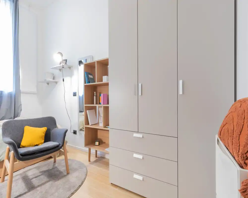 Rincón de dormitorio luminoso y moderno con armario, butaca y estantería; textiles suaves y detalles cálidos crean un ambiente acogedor.