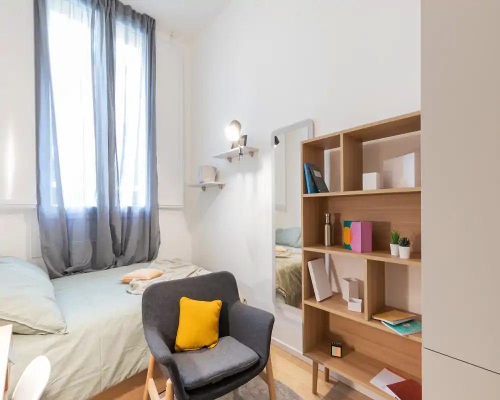 Dormitorio luminoso y compacto con cama bien hecha junto a una ventana alta, un sillón acogedor con cojín amarillo y una estantería de madera—bien presentado e ideal para estancias cortas.