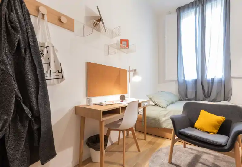 Dormitorio individual luminoso y bien decorado con escritorio de madera, lámpara de mesa y cama cómoda junto a una gran ventana; textiles suaves y un sillón acogedor crean un espacio ideal para trabajar y descansar.