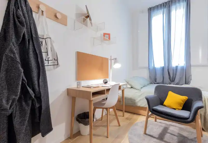 Habitación individual ordenada con escritorio, sillón y cama bien hecha bajo suave luz natural — acogedora, moderna y práctica para un estudiante o profesional.