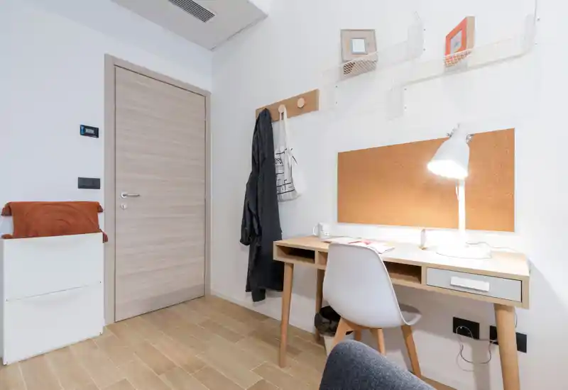 Compact bedroom corner staged with a simple wooden desk, cork noticeboard and a task lamp; neutral tones and warm wooden flooring create a functional, minimal space.