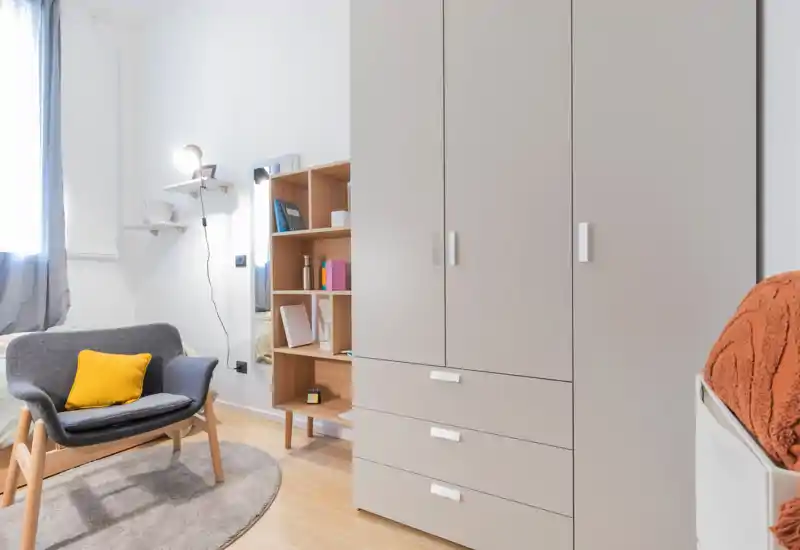 Small, modern bedroom corner with a grey armchair, yellow cushion, wardrobe and visible bed edge; bright, well-styled and cozy.