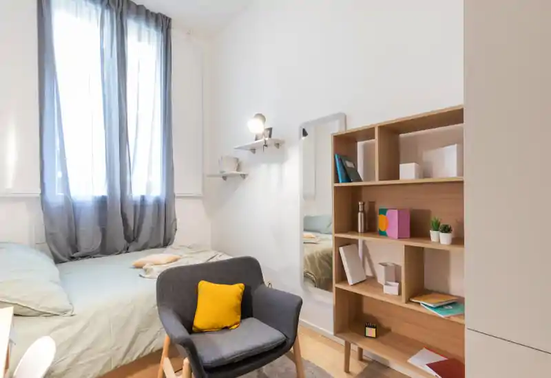 Bright, cozy single bedroom with a neatly made bed beside a large curtained window, a comfortable armchair with a yellow cushion, a full-length mirror and wooden shelving for storage.